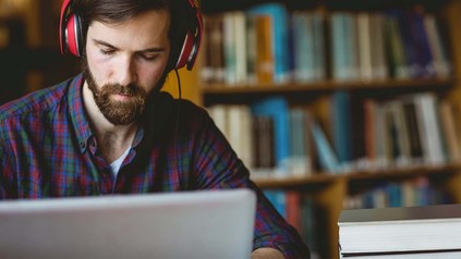 Bärtiger Student mit Kopfhörer arbeitet an Sitzplatz in Bibliothek.