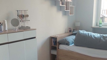 Ausschnitt aus einem Wohnheimzimmer mit Bett, Kommode, und Wandregal.