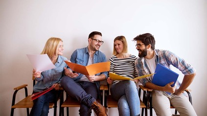 Vier Studierende mit Mappen voll mit Unterlagen auf Stühlen.