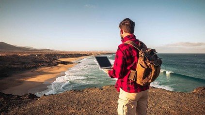 Mann in Karohemd an Strand mit Rucksack und Laptop.