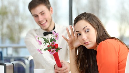 Ein Mann in weißem Hemd überreicht einer Frau in orangenem Pulli Blumen beim Date. Die Frau wendet sich mit entgeistertem Blick ab.