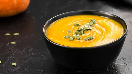 Kürbissuppe mit Kürbiskernen angerichtet auf einem Tisch, daneben der Teil eines Kürbis.