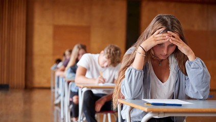 Tischreihe in Vorlesungssaal bei schriftlicher Prüfung, ganz vorne Studentin, die den Kopf auf ihre Hände abgestützt hat und auf das Blatt vor sich starrt.