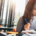 Junge Frau in grauem Pullover, die in der Bibliothek an einem Sitzplatz am Fenster arbeitet.