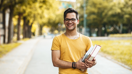 Junger Mann mit Brille und Büchern auf dem Arm, der auf dem Campus steht und lächelt.