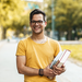 Junger Mann mit Brille und Büchern auf dem Arm, der auf dem Campus steht und lächelt.