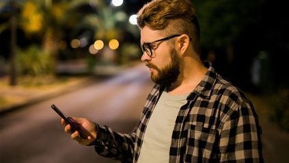 Junger Mann mit Karohemd und Brille, der nachts auf der Straße steht mit seinem Handy in der Hand.