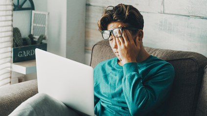 Junger Mann daheim auf dem Sofa mit Laptop auf dem Schoß.