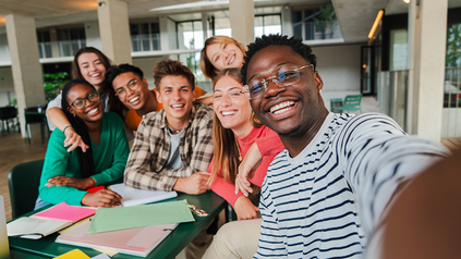 7 junge Leute um einen Tisch an der Uni - nehmen zusammen ein Selfie auf.