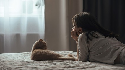 Junge Frau liegt mit Katze auf einem Bett liegend, in einem dunklen Zimmer.