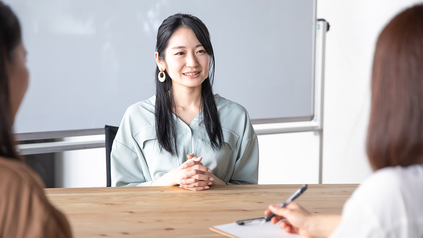 Asiatische Frau an Schreibtisch vor zwei Prüferinnen, eine davon macht sich Notizen.