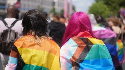 Zwei junge Menschen mit der Fahne der LGBTQIA+ Community um die Schultern von Hinten auf einer Demonstration.